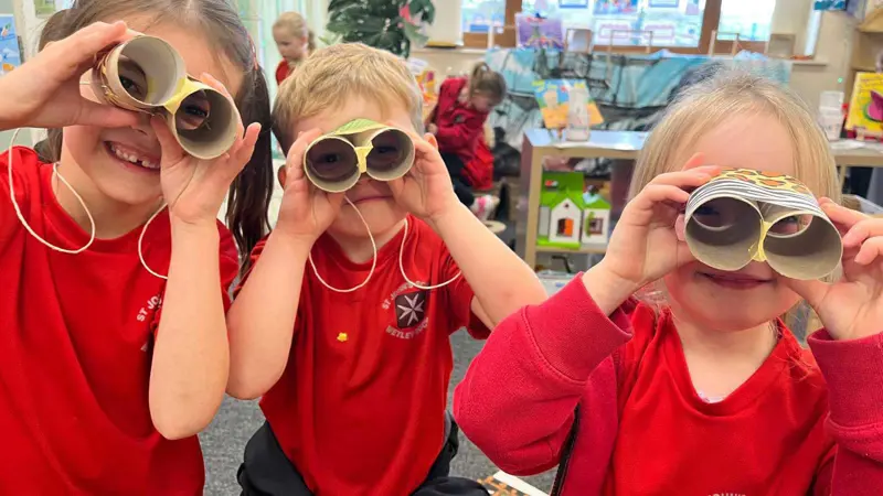 Three children smiling whilst looking at the camera through their binoculars.