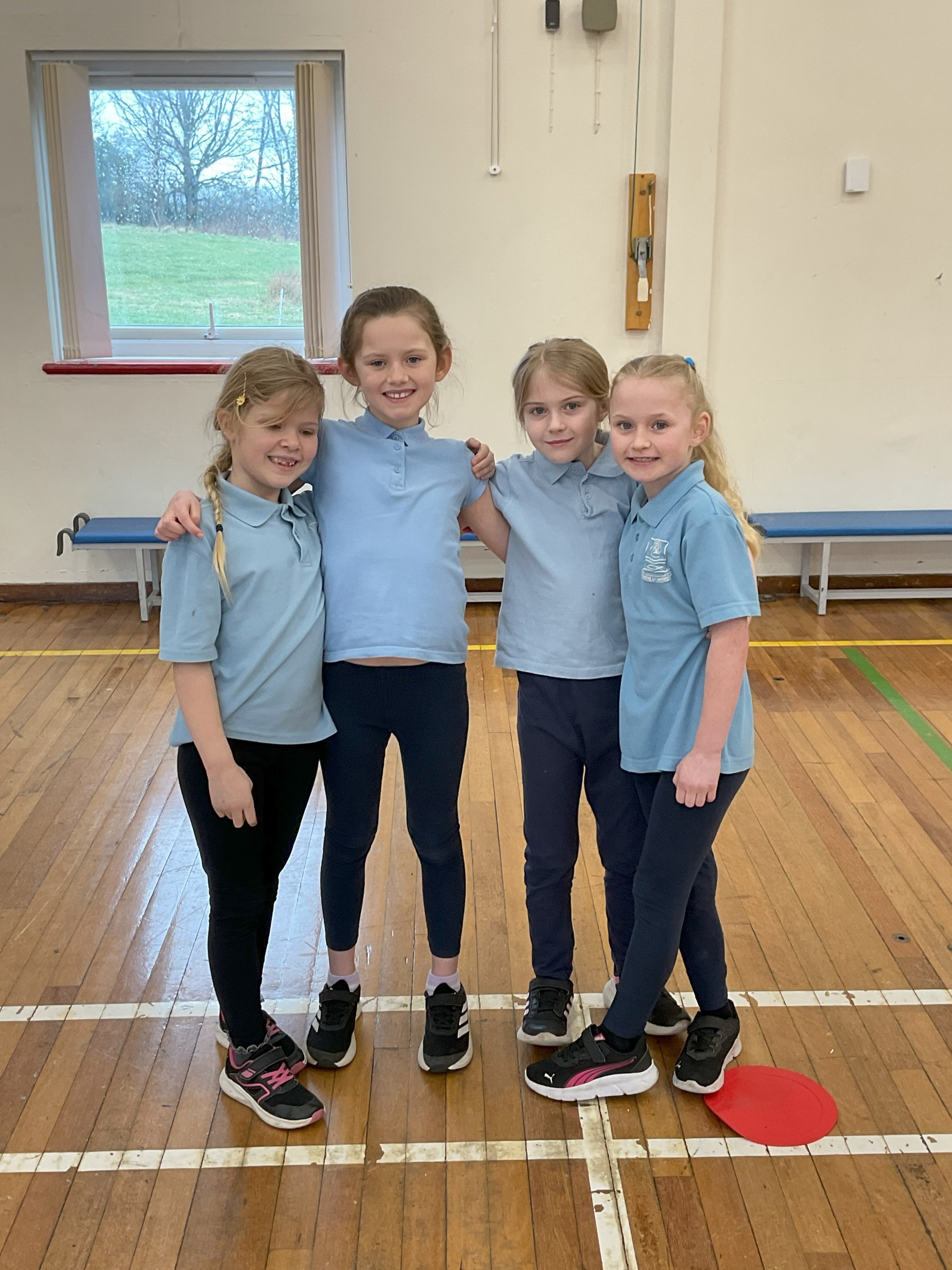 Four children smiling at a sporting event in a school hall.