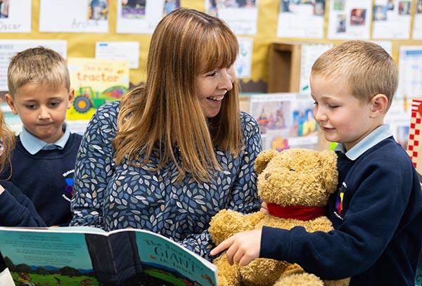 Teacher smiling with a child and teddy bear whilst sharing a book.