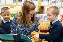 Teacher smiling with a child and teddy bear whilst sharing a book.